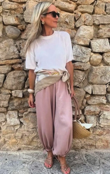 Uma mulher em pé contra uma parede de pedras, vestindo uma camiseta branca e calças largas cor de rosa. Ela usa acessórios como brincos grandes, uma faixa na cintura e óculos escuros, segurando uma bolsa de palha.