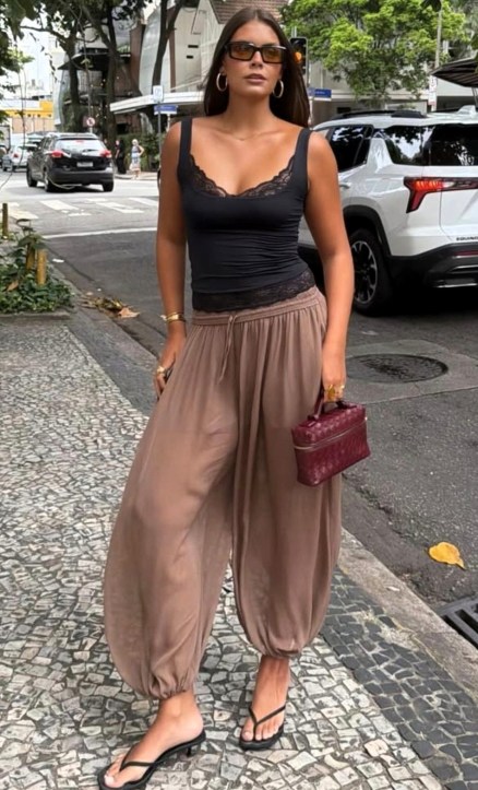 Mulher caminhando na rua com blusa preta e calças largas beje, segurando uma bolsa vermelha.
