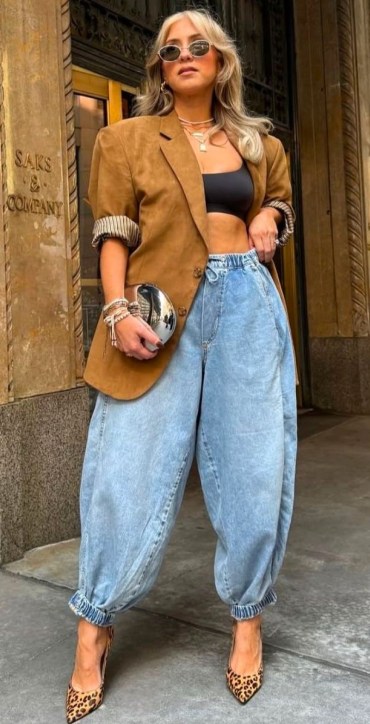 Mulher posando com um blazer marrom, top preto e calças jeans largas. Ela usa óculos de sol e sapatos de salto de onça.