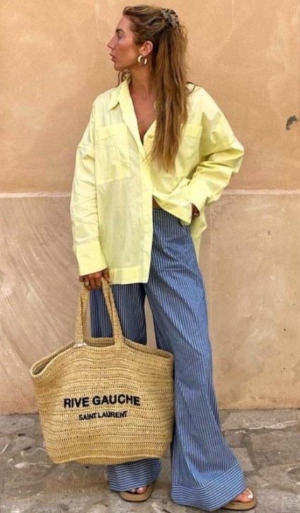 Mulher em um ambiente ao ar livre, usando uma camisa amarela oversized e calças listradas azuis. Ela segura uma bolsa de palha com o texto 'RIVE GAUCHE SAINT LAURENT' e tem o cabelo solto.