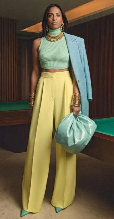 Mulher em pé, usando um top verde claro e calças largas amarelas, com um blazer azul claro e acessórios de ouro, segurando uma bolsa azul clara.