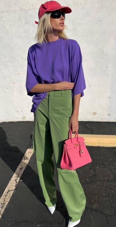 Modelo usando camiseta roxa, calça verde e bolsa rosa, com boné vermelho e óculos escuros, em um ambiente externo.