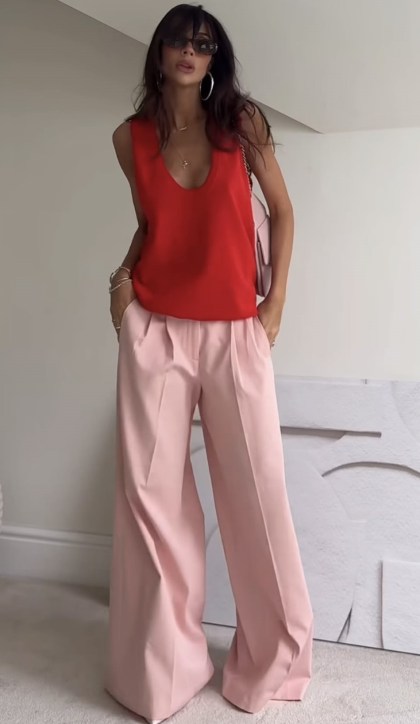 Mulher posando com blusa vermelha e calças largas rosa, em um ambiente interno com decoração minimalista.