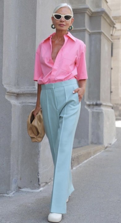 Mulher elegante usando camisa rosa e calças azul claro, segurando uma bolsa bege, com acessórios de moda e óculos escuros.