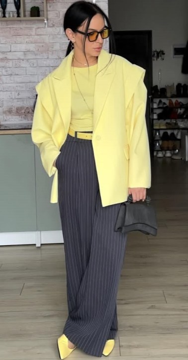Mulher usando um blazer amarelo, blusa verde e calças listradas em tons escuros. Ela segura uma bolsa preta e usa sapatos amarelos.