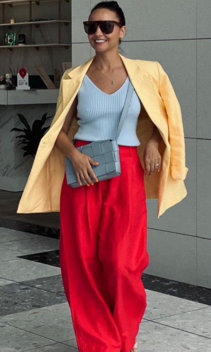 Pessoa sorrindo, usando um blazer amarelo, blusa azul e calça vermelha, segurando uma bolsa cinza, em ambiente interno moderno.