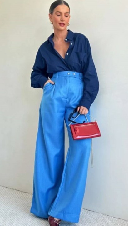 Mulher vestindo camisa azul-marinho e calças largas azuis, segurando uma bolsa vermelha.