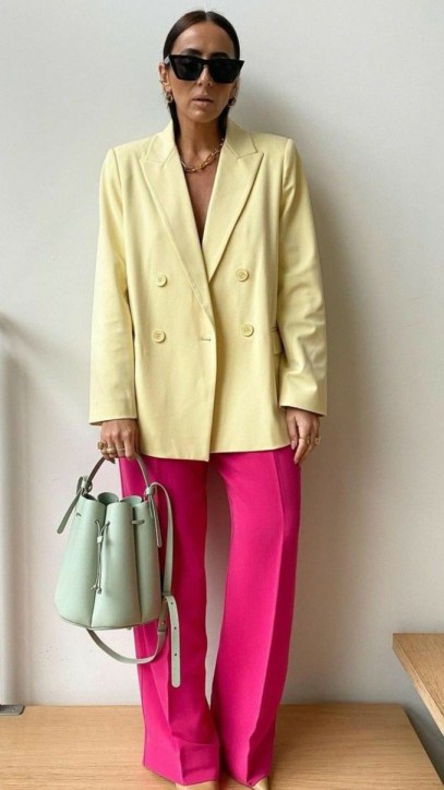 Mulher usando um blazer amarelo claro e calças rosa brilhante, com óculos escuros e bolsa verde-claro.