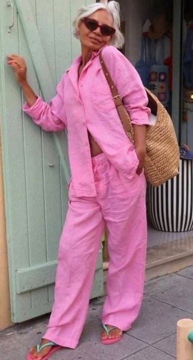 Mulher com cabelo branco, usando um conjunto rosa, posando ao lado de uma porta verde. Ela carrega uma bolsa de palha e está usando óculos escuros.