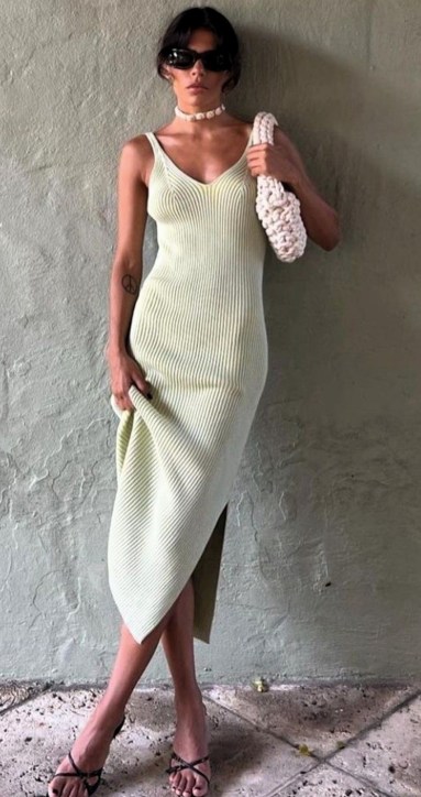 Uma mulher posa em frente a uma parede, vestindo um vestido midi de malha na cor verde claro, com um corte lateral, sandálias pretas e óculos escuros. Ela segura uma bolsa de crochê branca e usa um colar de pérolas.