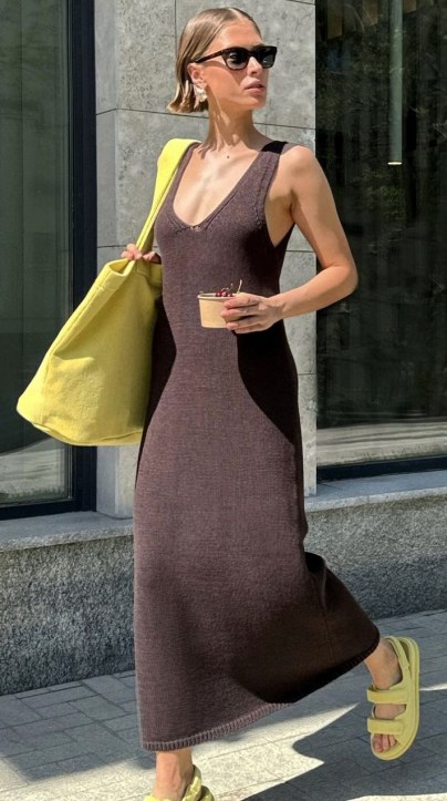 Mulher caminhando na calçada, usando um vestido longo marrom, óculos de sol e sandálias amarelas. Ela carrega uma bolsa amarela e um copo com bebida.
