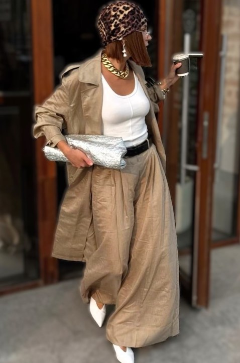 Mulher caminhando com uma jaqueta bege, blusa branca e saia longa, segurando uma bolsa prateada e um copo de bebida.