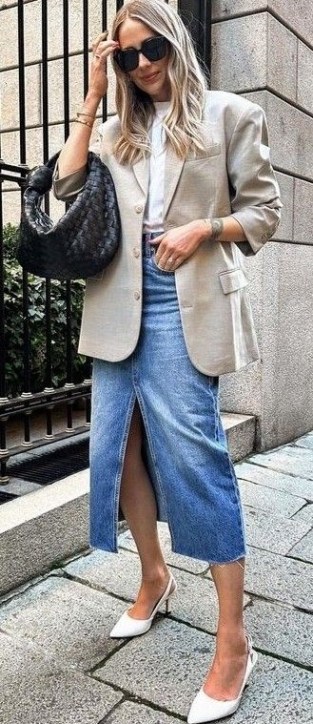 Mulher loira usando um blazer claro, camiseta branca, saia jeans com fenda e sapatos de salto alto brancos, segurando uma bolsa preta.