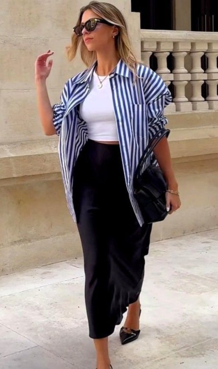 Mulher caminhando em uma rua, vestindo uma camisa listrada azul e branca e uma saia longa preta, com óculos escuros e um bolsa preta.