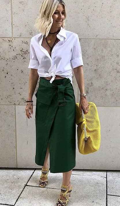 Mulher vestindo uma camisa branca amarrada na cintura e uma saia verde, segurando uma bolsa amarela e usando sandálias com tiras.