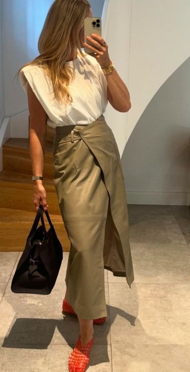 Mulher usando blusa branca e saia longa cinza, segurando uma bolsa preta e utilizando sapatos vermelhos.