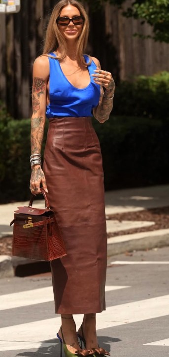 Uma mulher sorridente com cabelo longo e tatuagens, vestindo uma blusa azul e uma saia longa de couro marrom, segurando uma bolsa vermelha enquanto caminha na rua.