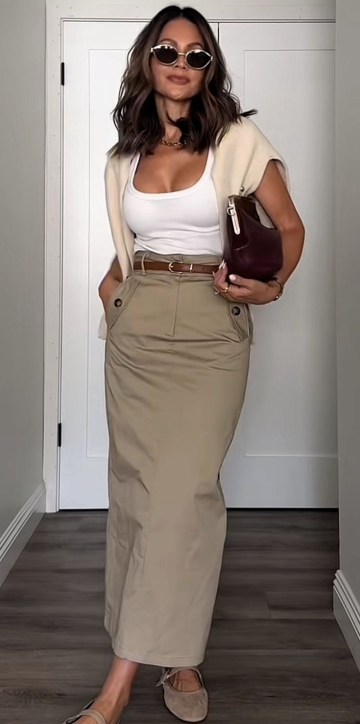 Mulher de pé em um ambiente interno, usando uma blusa branca e uma saia longa bege. Ela está com óculos de sol, um colete leve e segurando uma bolsa marrom, aparentando um estilo casual e elegante.
