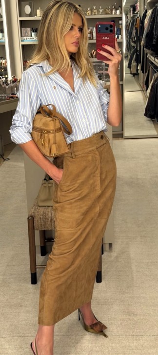 Mulher posando para um autorretrato, vestindo uma camisa de listras azuis e brancas e uma saia longa marrom. Ela segura uma pequena mochila e está em um ambiente de moda com prateleiras ao fundo.