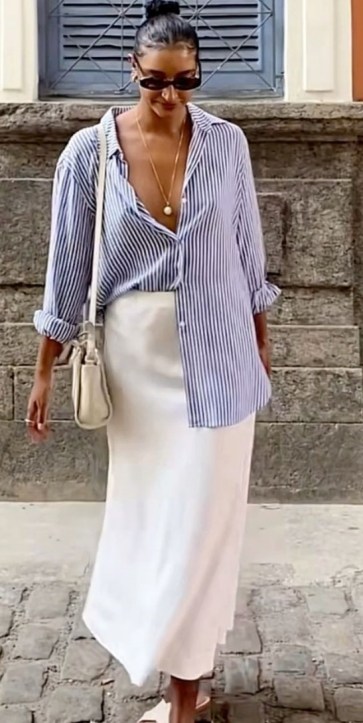 Mulher usando uma camisa listrada azul e branca e uma saia branca, caminhando em uma calçada de pedra.